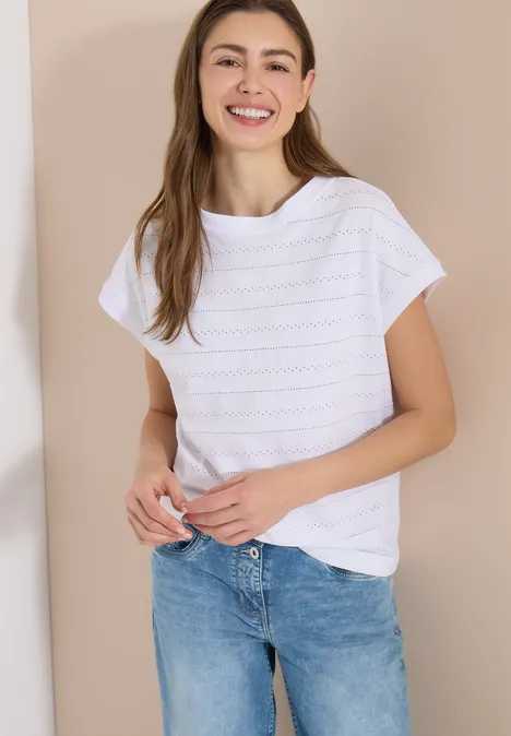 Frau mit weißem T-Shirt und blauen Jeans lehnt an hellem Hintergrund.