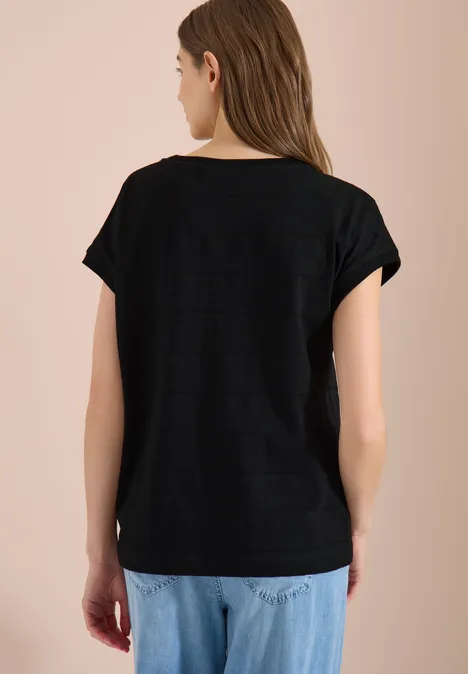 Frau steht mit dem Rücken zur Kamera, trägt schwarzes T-Shirt und helle, lockere Jeans vor beigem Hintergrund.