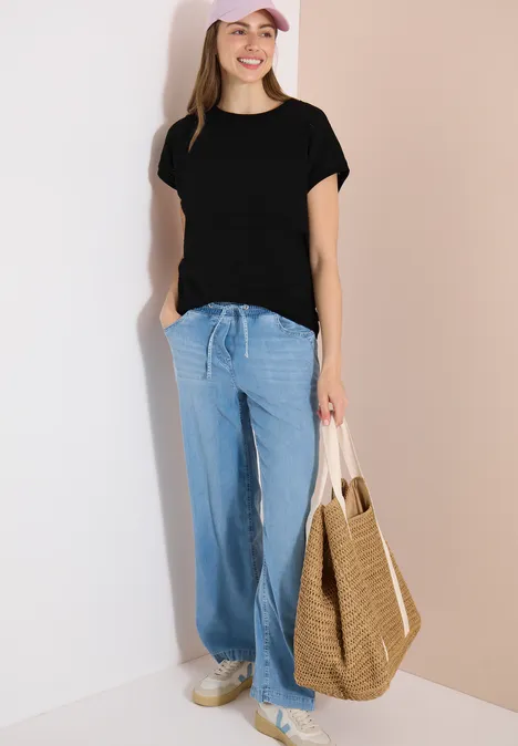 Frau im schwarzen T-Shirt, weiten hellblauen Hosen und weißen Sneakern, mit geflochtener Strohtasche.
