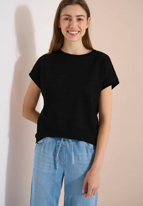 Person trägt einfaches schwarzes T-Shirt und lockere hellblaue Hosen vor neutralem Hintergrund.