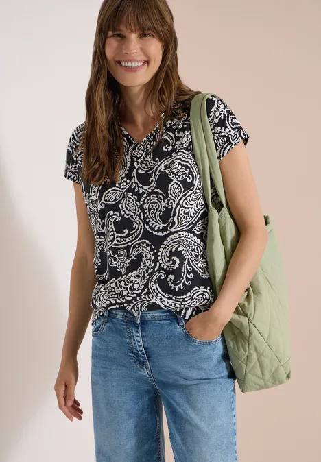 T-Shirt mit V-Neck und Print Black Frau in gemustertem T-Shirt, blauen Jeans, grüner Stepp-Tasche, lässig mit Hand in der Hosentasche.