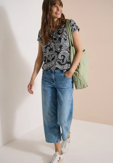 T-Shirt mit V-Neck und Print Black Frau trägt gemustertes T-Shirt, weite Cropped-Jeans, weiße Sneaker und einen grünen Steppbeutel.