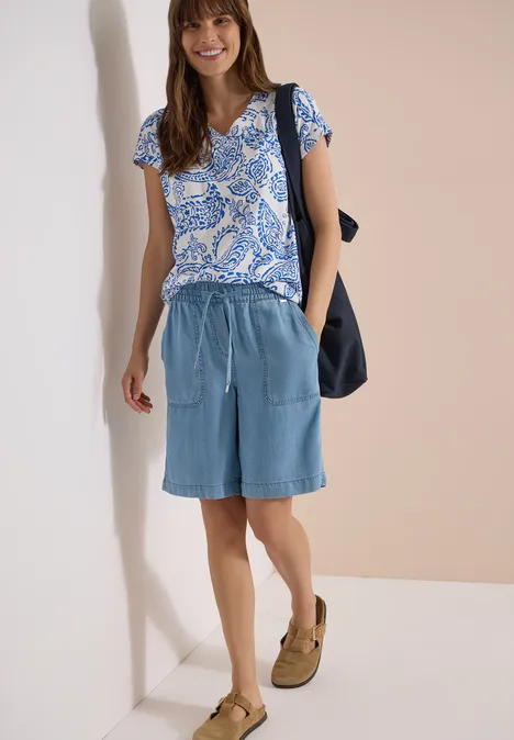 T-Shirt mit V-Neck und Print vanilla white Frau mit blauem gemustertem T-Shirt, hellblauen Jeansshorts, Sandalen und schwarzer Tasche vor heller Wand