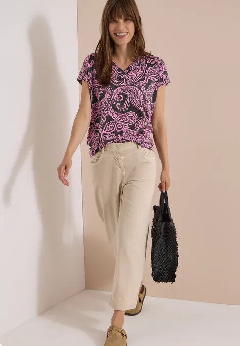 T-Shirt mit V-Neck und Print raw khaki Frau trägt lila Paisley-Shirt, beige Hose und schwarze Tasche.