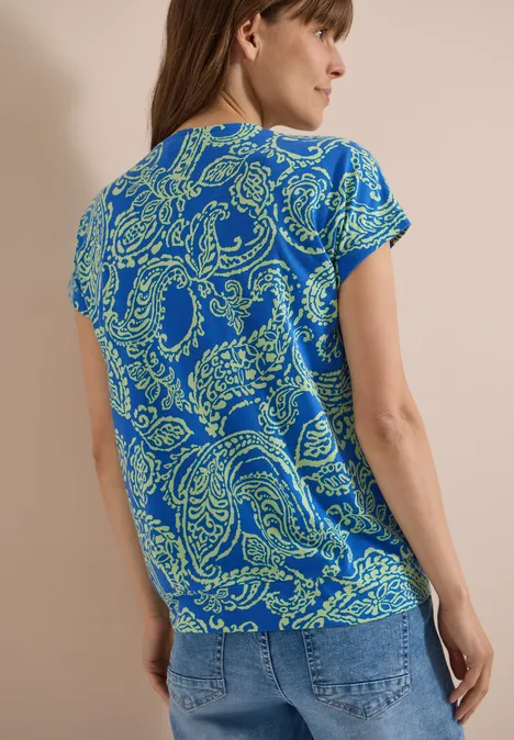 Frau von hinten mit blauem T-Shirt und grünem Paisleymuster, dazu Jeans.