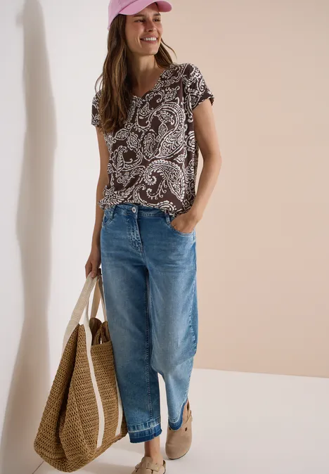 T-Shirt mit V-Neck und Print brown cacao Frau in gemustertem Top, blauen Jeans und Basttasche, lässiger Look mit Loafers.