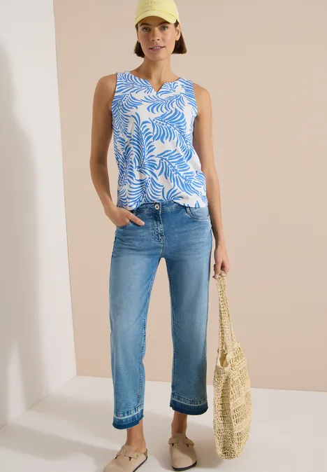 Femme en jean clair, top sans manches bleu à motifs feuilles, sac en paille, fond neutre.