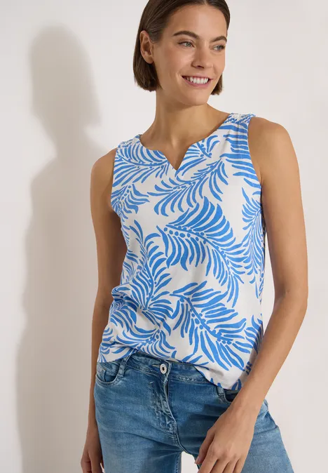 Femme portant un top sans manches à motif de feuilles bleues et un jean sur fond blanc.