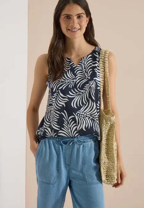 Top met split in de hals en tweekleurig patroon universal blue Vrouw in donkerblauwe top met bladprint, lichtblauwe broek en gevlochten strandtas