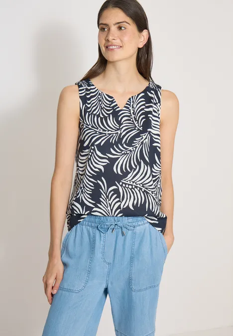 Top met split in de hals en tweekleurig patroon universal blue Vrouw draagt een mouwloos marineblauw topje met wit bladerenpatroon en een lichte jeansbroek.