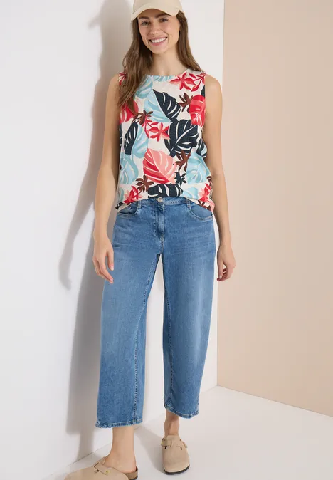 Top mit Rundhals und Blätterprint light alabaster white Frau in floralem ärmellosem Top, weiten blauen Culotte-Jeans und beigefarbenen Slippern vor hellem Hintergrund