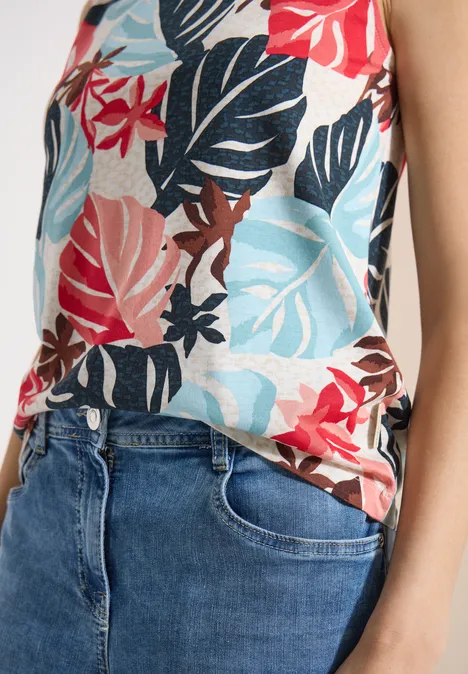 Top mit Rundhals und Blätterprint light alabaster white Frau trägt ärmelloses Top mit buntem Blattmuster und blauen Jeans, Hände in den Hosentaschen.