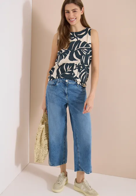 Top met ronde hals en bladprint palm breeze green Vrouw draagt mouwloze top met bladprint, wijde cropped jeans, lichte sneakers en gele strooien tas.