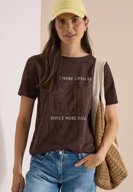 T-Shirt in Ausbrenner-Optik brown cacao Junge Frau trägt braunes T-Shirt mit Aufdruck, Jeans und Basttasche vor beigem Hintergrund.