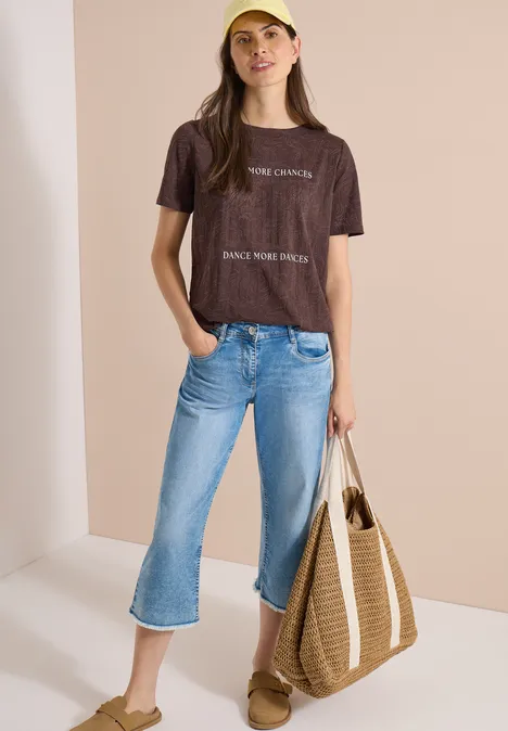 T-Shirt in Ausbrenner-Optik brown cacao Eine Person trägt braunes T-Shirt, blaue Cropped-Jeans, Sandalen und eine große Strohtasche vor neutralem Hintergrund.