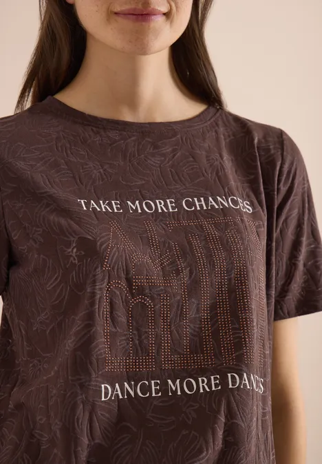 T-Shirt in Ausbrenner-Optik brown cacao Portrait einer Frau im braunen T-Shirt mit Mustern und den Slogans ‚TAKE MORE CHANCES‘ und ‚DANCE MORE DAYS‘.