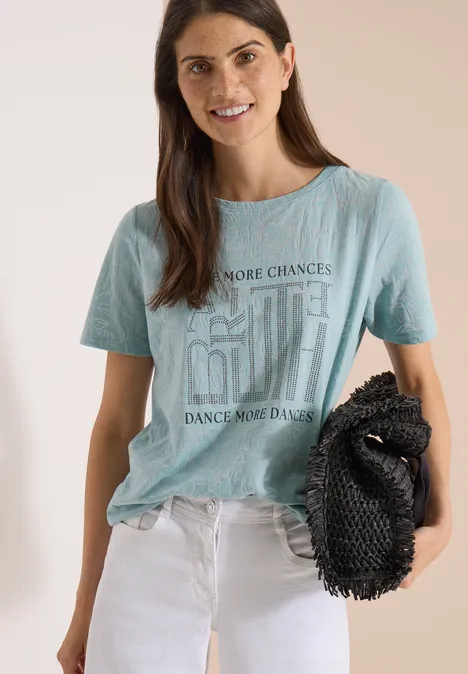 T-Shirt in Ausbrenner-Optik crystal mint Junge Frau trägt hellblaues T-Shirt mit Aufdruck, weiße Hose und schwarze Clutch, vor neutralem Hintergrund.