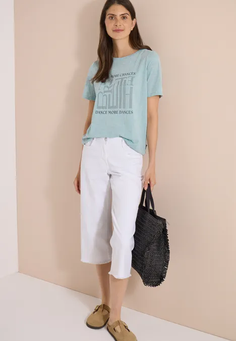 T-Shirt in Ausbrenner-Optik crystal mint Frau vor beige-eckiger Wand, trägt hellgrünes T-Shirt mit Print, weiße Caprihose, beige Schuhe, schwarze Tasche.