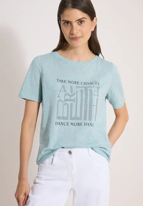 T-Shirt in Ausbrenner-Optik crystal mint Frau trägt hellgrünes T-Shirt mit Aufschrift und weiße Hose vor hellem Hintergrund.