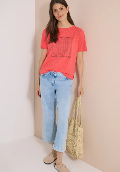 T-Shirt in Ausbrenner-Optik coral Frau in rotem T-Shirt, hellen Jeans, trägt Basttasche und Sandalen.