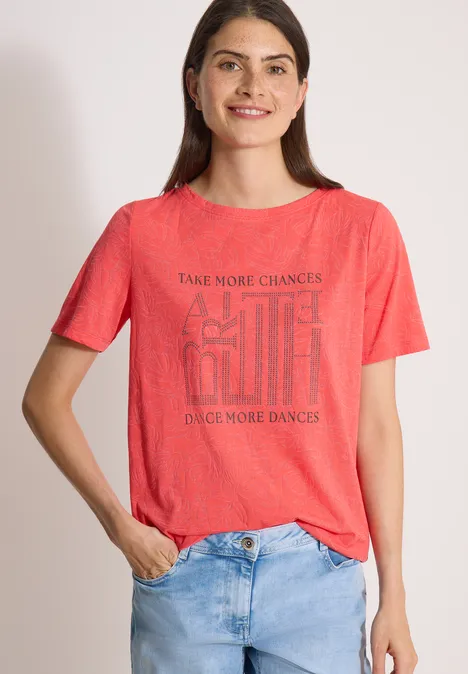 T-Shirt in Ausbrenner-Optik coral Eine Frau in rotem T-Shirt mit Schriftzug und hellblauen Jeans steht vor hellem Hintergrund.