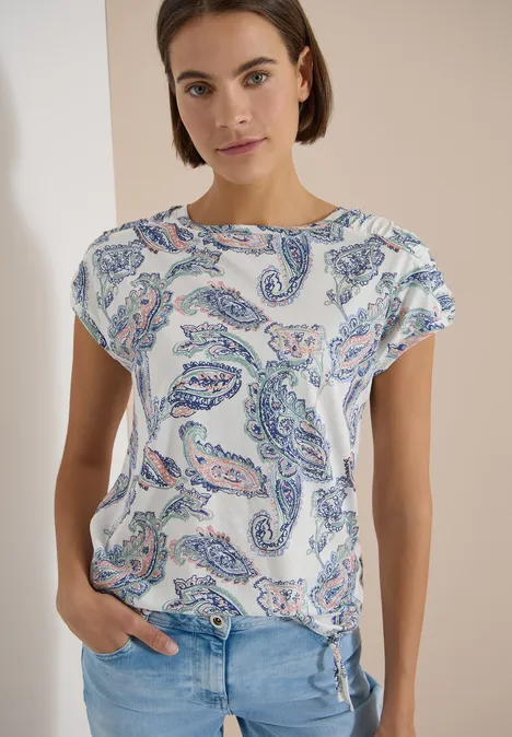 Eine Frau trägt ein weißes T-Shirt mit buntem Paisley-Muster und blaue Jeans vor hellem Hintergrund.