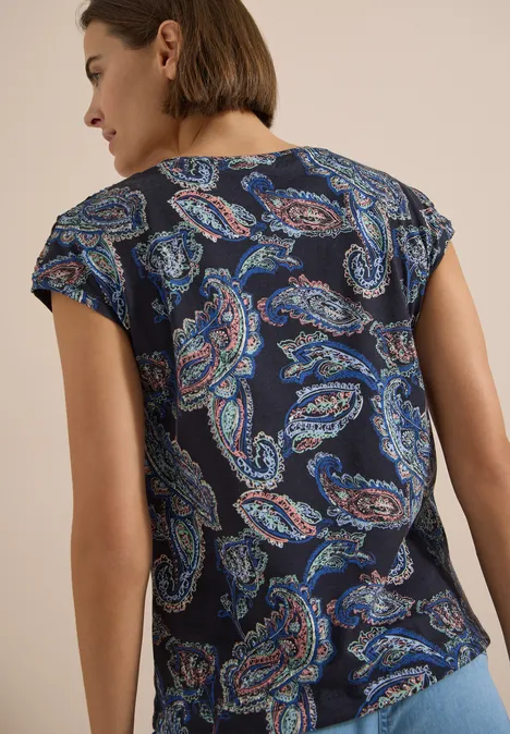 Vrouw van achteren in donkerblauw T-shirt met meerkleurig paisleymotief, lichte achtergrond.