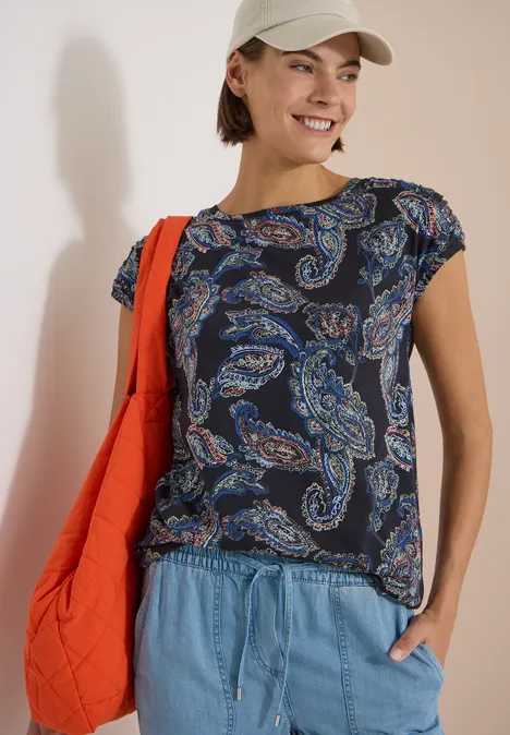 Vrouw met paisley-shirt, spijkerbroek en rode schoudertas.