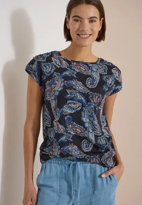 Vrouw in donkerblauw t-shirt met paisley-print en lichtblauwe jeans, nonchalant met handen in zakken.