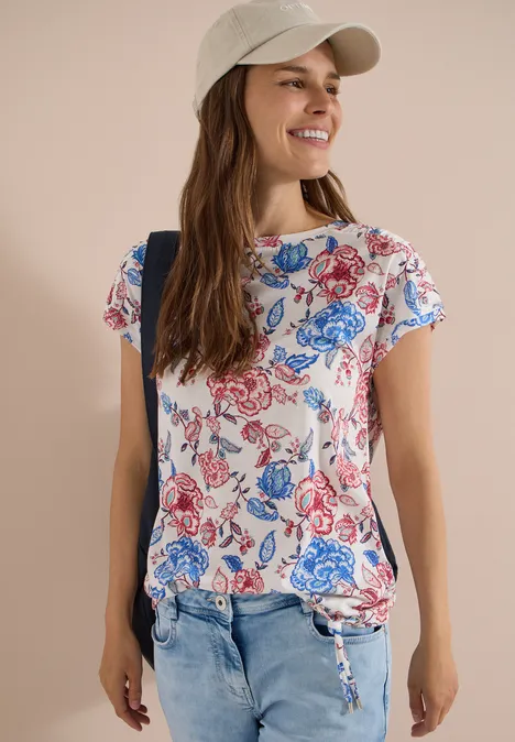 Kurzarm Shirt mit Boatneck und Raffung vanilla white Frau in Jeans, weißem T-Shirt mit blau-rotem Blumenprint und Baseball-Cap.