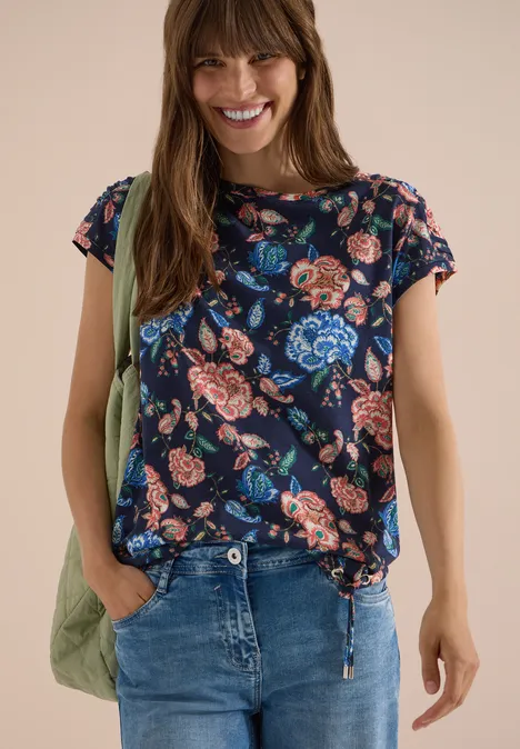 Kurzarm Shirt mit Boatneck und Raffung urban dark blue Frau in Jeans trägt ein dunkelblaues T-Shirt mit buntem Blumenmuster und eine grüne Umhängetasche.