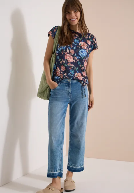 Kurzarm Shirt mit Boatneck und Raffung urban dark blue Frau in geblümtem Shirt, weiten Jeans, beigen Sandalen und grüner Tasche vor schlichtem Hintergrund.