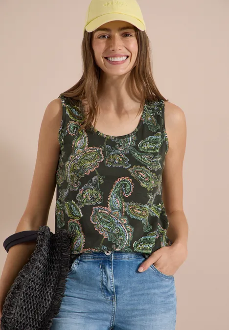 Top mit gerundetem V-Neck und Flechtdetail raw khaki Frau in ärmellosem Paisley-Top und blauen Jeans vor neutralem Hintergrund, hält eine Flechtasche.