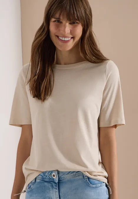 Eine Person trägt ein schlichtes beiges T-Shirt und blaue Jeans vor hellem Hintergrund.