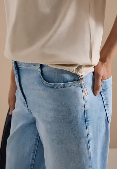 Seitliche Ansicht einer Person in hellblauen Jeans und weißem T-Shirt, Hand in Tasche, sichtbarer Schulterriemen.