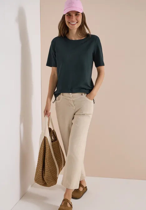 Dame vor beiger Wand in dunkelgrünem T-Shirt, hellen Cordhosen, mit Korbtasche und braunen Sandalen.