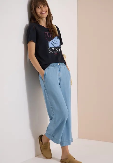 T-Shirt mit Rundhals und Frontprint universal blue Frau lehnt an Wand, trägt schwarzes T-Shirt mit Print, weite hellblaue Jeans und braune Mokassins.