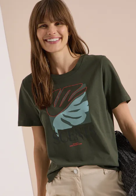 T-Shirt mit Rundhals und Frontprint khaki Person trägt ein khakigrünes T-Shirt mit Blättermotiv und eine beige Hose, hält einen dunklen Pullover in der Hand.