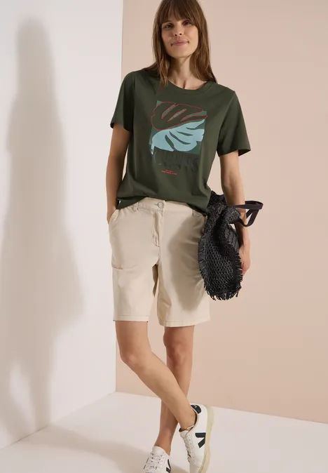 T-Shirt mit Rundhals und Frontprint khaki Person lehnt an Wand, trägt grünes T-Shirt mit Blattdesign, beiges Bermuda, weiße Sneaker und schwarzen Netzbeutel.