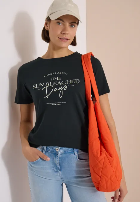 T-Shirt mit Boat Neck und Wording Artwork palm breeze green Person in schwarzem T-Shirt, hellblauen Jeans, orangefarbener Stepp-Tasche und Baseballcap vor weißer Wand.
