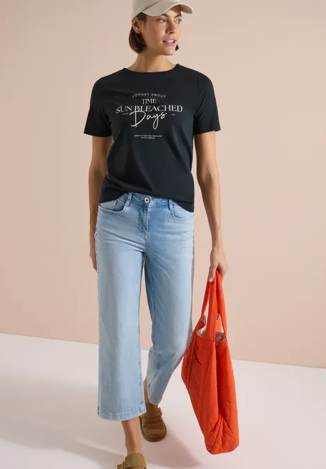 T-Shirt mit Boat Neck und Wording Artwork palm breeze green Frau in schwarzem T-Shirt, hellblauen Jeans und braunen Schuhen trägt orangefarbene Tasche vor beiger Wand.
