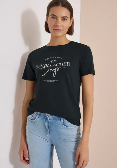 T-Shirt mit Boat Neck und Wording Artwork palm breeze green Eine Frau in Jeans und schwarzem T-Shirt mit weißem Aufdruck, vor hellem Hintergrund.