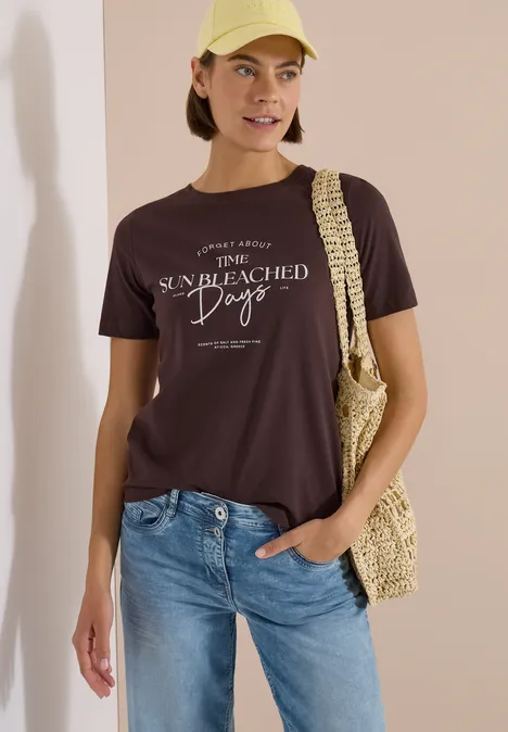 T-Shirt mit Boat Neck und Wording Artwork brown cacao Frau in blauen Jeans und dunkelbraunem T-Shirt mit weißem Print, trägt eine Strohtasche vor hellem Hintergrund.