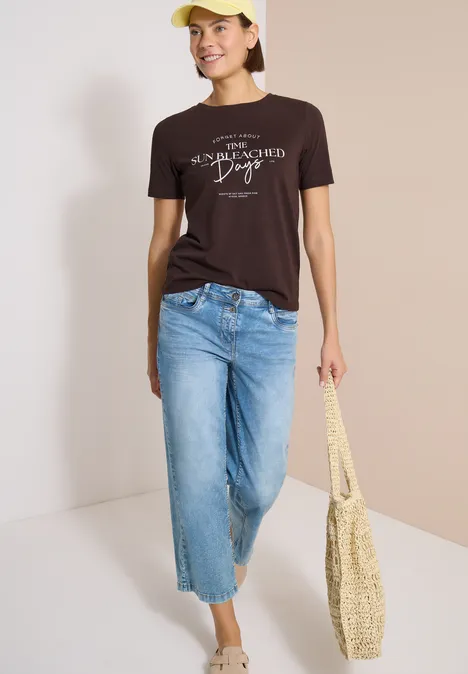 T-Shirt mit Boat Neck und Wording Artwork brown cacao Person mit braunem T-Shirt, hellen Jeans, gelber Mütze und Strohtasche vor hellem Hintergrund