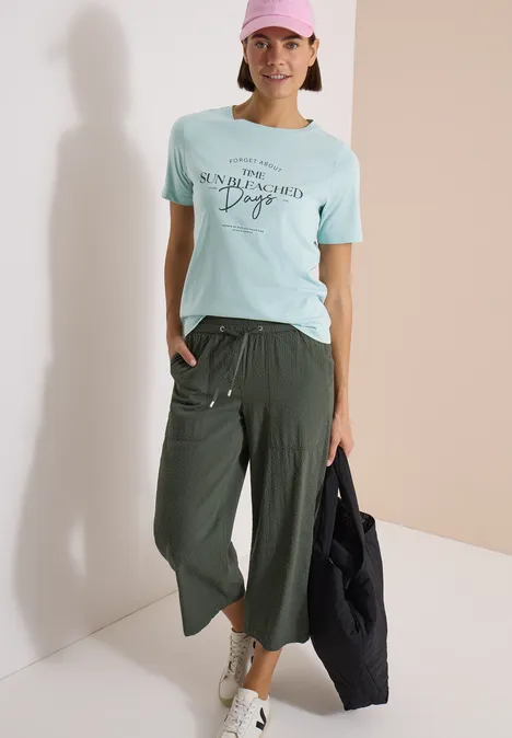 T-Shirt mit Boat Neck und Wording Artwork crystal mint Person trägt mintfarbenes T-Shirt mit Aufdruck, weite grüne Culotte, weiße Sneaker und schwarzen Shopper.