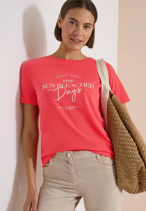 T-Shirt mit Boat Neck und Wording Artwork coral Frau trägt korallrotes T-Shirt mit weißem Aufdruck 'Sun Bleached Days', hellbeige Hose und Strandtasche.