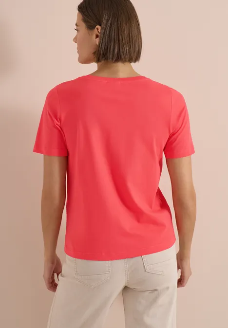 T-Shirt mit Boat Neck und Wording Artwork coral Frau von hinten in korallenrotem T-Shirt und beigen Hosen vor neutralem Hintergrund.