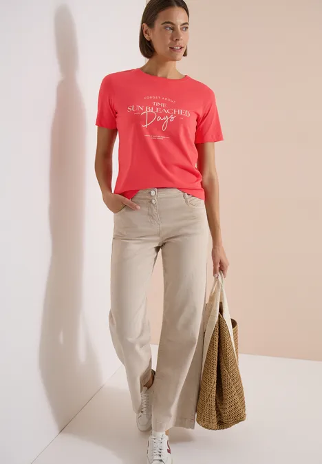 T-Shirt mit Boat Neck und Wording Artwork coral Frau in hellem Korall-T-Shirt, beiger Hose und weißen Sneakern mit großer Basttasche.