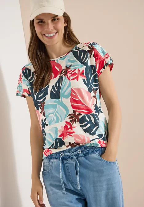 Vrouw draagt kleurrijk t-shirt met tropische bloemenprint en lichtgewicht jeans met koordsluiting.