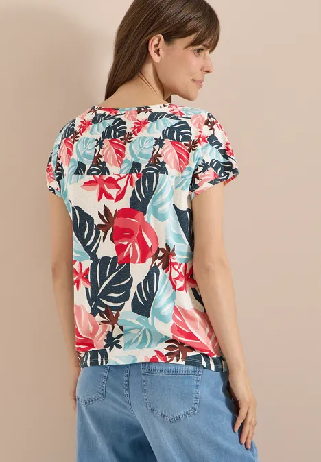 Vrouw van achteren in een shirt met korte mouwen met kleurrijk bladerenpatroon (rood, turkoois) en lichtblauwe jeans.